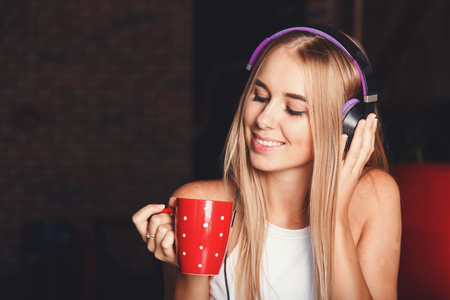 Cute blonde long hair girl wearing sleapwear sitting in the red loft comfortable sunny kitchen, listening music and drinking her morning coffeeの写真素材