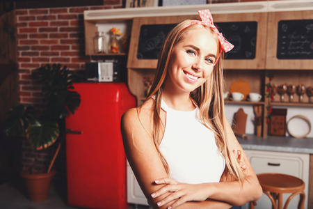 Nice blonde long hair housewife smiling in the red loft comfortable sunny kitchen and preparing for the cookingの写真素材