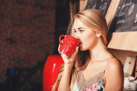 Lovely blonde long hair girl wearing sleapwear standing in the red loft comfortable sunny kitchen, drinking coffee and ready to the new dayの写真素材