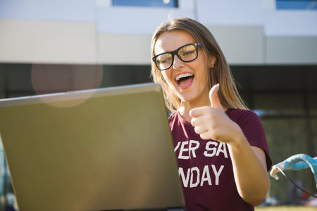Happy hipster student woman wearing glasses showing thumb near the university and rejoicing good exam results, she sitting with open laptop, studying conceptionの写真素材