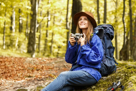 Young pretty woman with backpack and camera wearing hat hiking in autumn forest in mountains, she...