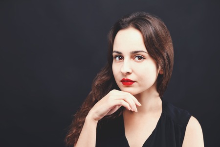 Portrait of pretty serious brunette woman wearing black dress standing on the black background,...