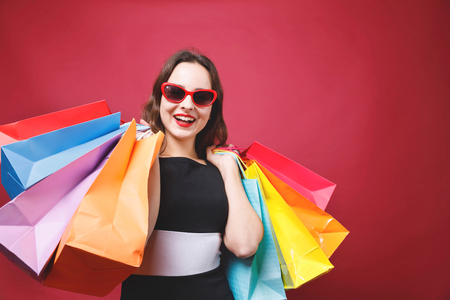 Young cute girl with bright makeup in black dress holding up many colorful paper shopping bags and looking at camera with smiling face on red backgroundの写真素材
