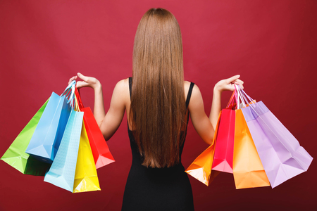 View of young attractive long hair woman in black dress standing in studio with paper shopping bags on red background, back viewの写真素材