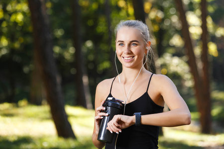 Athletic long-haired girl wearing black shirt standing in the sunny park, listening music and looking on the sports watch, she ready for the morning runningの写真素材