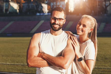 Portrait of multicultural handsome bearded man in blue shirt and sporty pretty blonde long haired woman in white t-shirt at the football stadium outdoor, green grass backgroundの写真素材