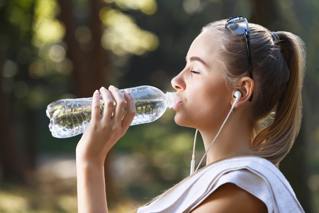 Young athletic woman with towel standing in summer park while drinking water after exerciseの写真素材