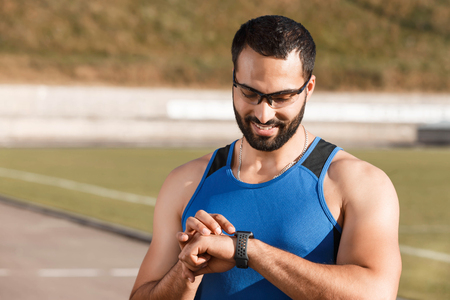 Athletic multicultural bearded man wearing sporty glasses smiling while looking on the indicators on his sport watch on the stadiumの写真素材