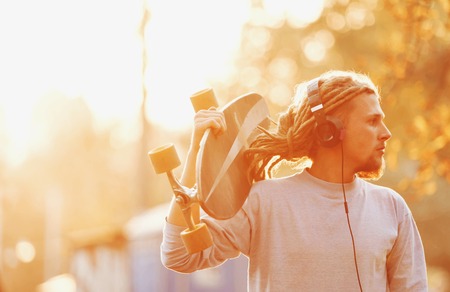 Hipster skater blonde bearded man with dreads wearing headphones walking in the forest and looking on the sunset, outdoor viewの写真素材