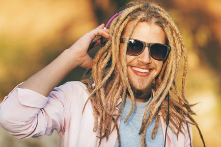 Portrait of hipster blonde bearded boy with dreads wearing headphones and glasses walking, smiling and listening his favorite music outdoors an the sunny autumn dayの写真素材