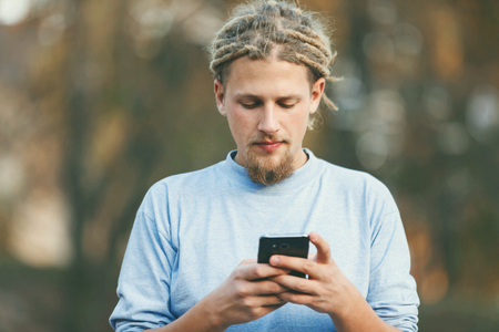 Hipster blonde bearded man with dreads wearing casual clothes watching movie on his smartphone, outdoor viewの写真素材