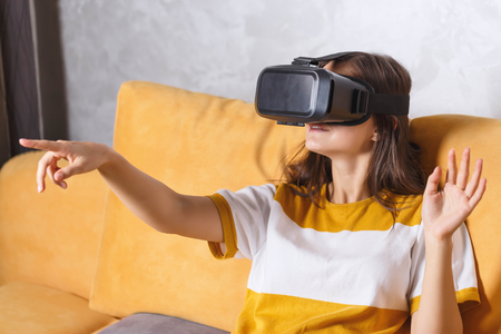 Attractive long-haired girl in pullover gesturing while testing VR device, she sitting on the yellow sofa in the light living room, future technology conceptの写真素材