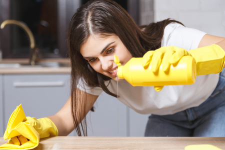Pretty long-haired girl in white t-shirt wearing yellow rubber gloves pours liquid on the table on the kitchenの写真素材