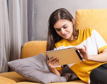 Cute long haired brunette girl laying on the yellow sofa in the light living room and browsing Internet on the digital tablet, modern technology, communication conceptの写真素材