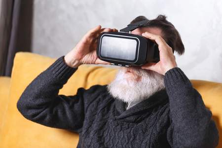 Contemporary senior bearded man in grey pullover wearing a virtual reality headset while sitting on the yellow sofa in the light living room, future technology conceptの写真素材