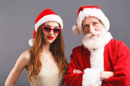 Santa Claus and young mrs. Claus wearing Santa hat and sunglasses standing on the gray background, New Year, Christmas, holidays, souvenirs, gifts, shopping, discounts, shops, Snow Maiden Santa Claus,make-up, hairstyle, carnival.の写真素材