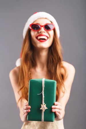 Hilarious cute woman in red santa claus hat and sunglasses with red lips holding present on outstretched arms, she laughing and standing on the gray background, New Year, Christmas, holidays, souvenirs, gifts, shopping, discounts, shops, Snow Maiden Santa Claus,make-up, hairstyle, carnival.の写真素材