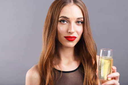 Portrait of pretty long haired woman with red lips wearing black dress standing on the gray background while drinking champagne, New Year, Christmas, holidays, souvenirs, gifts, shopping, discounts, shops, make-up, hairstyle, carnival.の写真素材