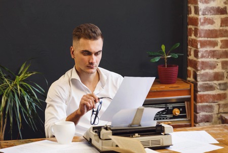 Young writer wearing glasses and white shirt sitting near the typewriter and looking critically on...