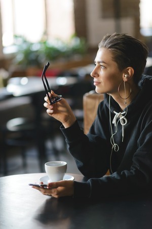 Pensive shortcut hipster girl holding her glasses and looking to the window while drinking coffee at the cafeteriaの写真素材