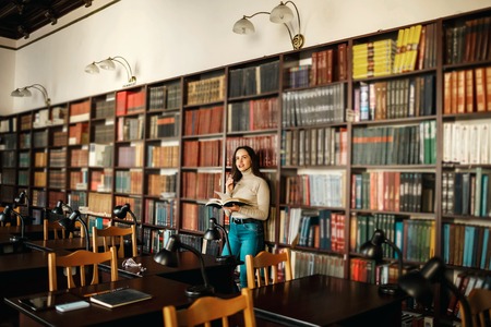 Young attractive student girl reading a book between library bookshelvesの写真素材