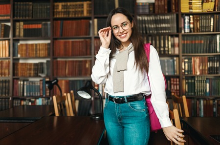 Brunette student girl in the white shirt wearing glasses standing with pink backpack at the libraryの写真素材