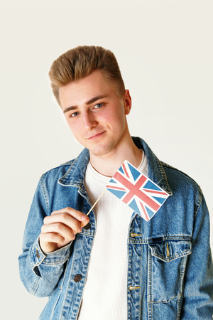 Teen blonde man wearing white t-shirt and jeans jacket holding small flag of England while standing on the white backgroundの写真素材