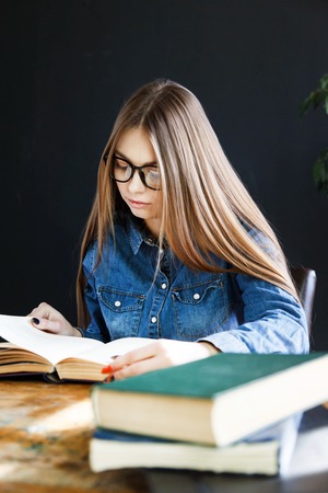 Student long haired girl wearing glasses sitting by wooden table and reading bookの写真素材