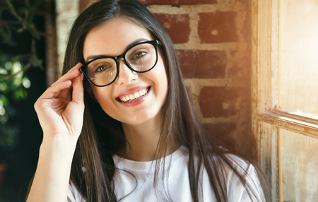 Brunette longhaired woman holding her glasses and smiling near the windowの写真素材