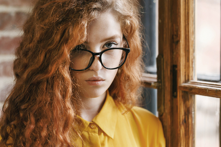 Portrait of pensive curly ginger girl in the yellow shirt wearing glasses standing near the window at the loft placement and looking seriously to the camera.の写真素材