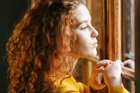 Teen curly ginger girl in the yellow shirt looking to the big window at the loft interiorの写真素材