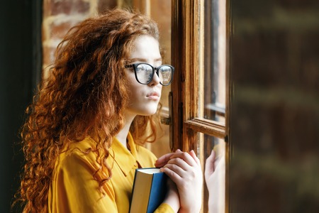 Portrait of curly ginger girl in the yellow shirt wearing glasses holding book and looking to the...