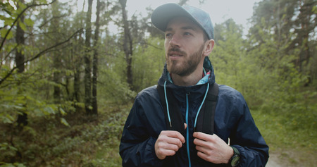 Bearded man dressed in cap and backpack walking in the forest