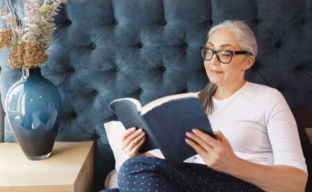 Pretty old woman with grey hair in eyeglasses reading book in bedroom