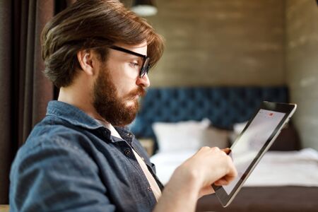 Bearded hipster man wearing glasses using tablet at the bedroom
