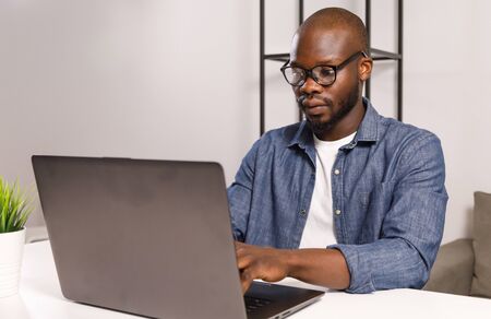 Serious african man wearing glasses watching and working on computer laptop at homeの写真素材
