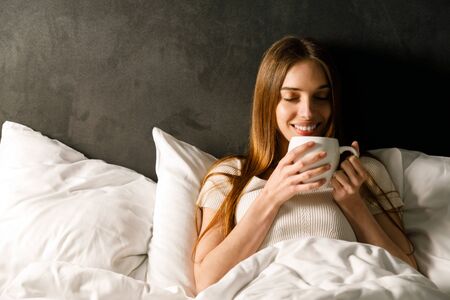 Young woman drinking tea in bed, enjoying morning in soft lightの写真素材