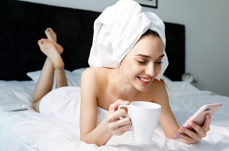Beautiful woman with towel on head typing message and drinking moning coffee in bedroom at homeの写真素材