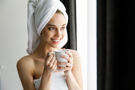 Young beautiful smiling woman dreaming while standing near the window and holding a cup of tea or coffeeの写真素材