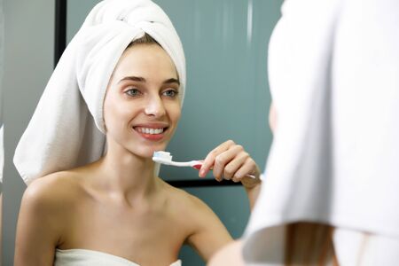 Portrait of a beautiful woman in bath towel brushing teeth and looking in the mirror in the bathroom