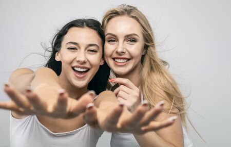 Joyful cute girlfriends in white tank tops smiling and stretching hands to camera isolated white backgroundの写真素材