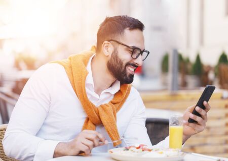 Bearded male freelancer working remotely on cafe terrace talking on mobile phone while eatingの写真素材