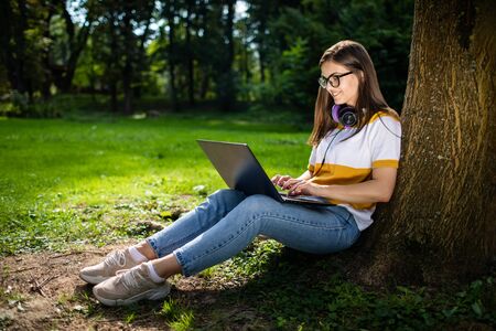 Smiling cute girl using a laptop resting against a park tree
