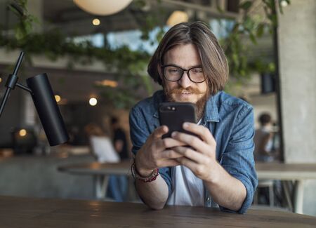 Bearded brown-haired young man using smartphone in a loft cafe