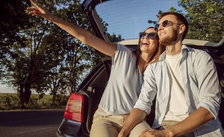 Young cheerful couple admires the countryside views while sitting in the car trunk in the rays of sunset sunの写真素材