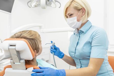 Female pediatric dentist with smiling eyes using water spray syringe to cleanse young patient's...