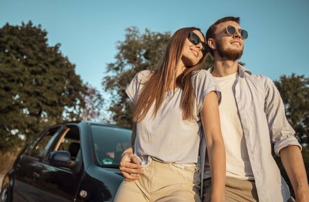 Loving couple posing on a sunset countryside road leaning against car hood low angle viewの写真素材