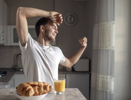 Young man stretches himself in a sunny kitchen with tasty croissants and orange juice waiting for himの写真素材