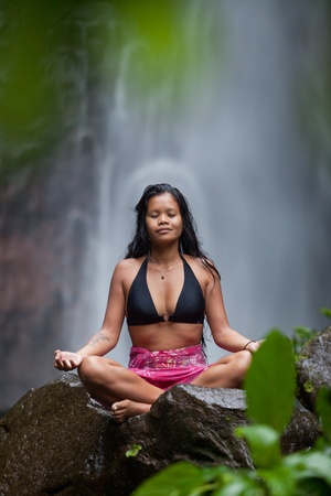 Woman sitting in meditation on rock at waterfall in tropical rainforestの写真素材