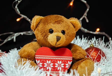 A brown teddy bear sits with a Christmas tree toy in the form of a heart on white tinsel, surrounded by red Christmas tree decorations. In the background is a black background with a garlandの写真素材